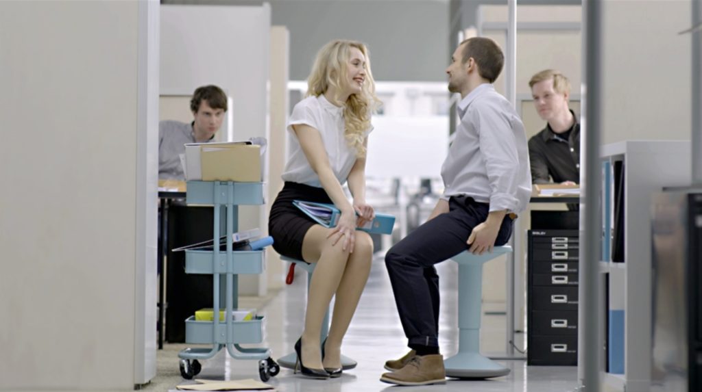 Up Stool man and woman chatting while seated on blue Up Stools in cubicle environment