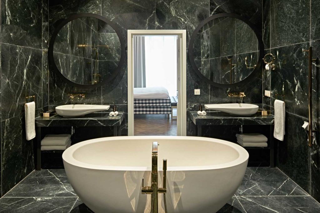 Hotel PACAI bathroom with green marble walls, dual sinks, and oval tub in center of frame with bedroom visible beyond