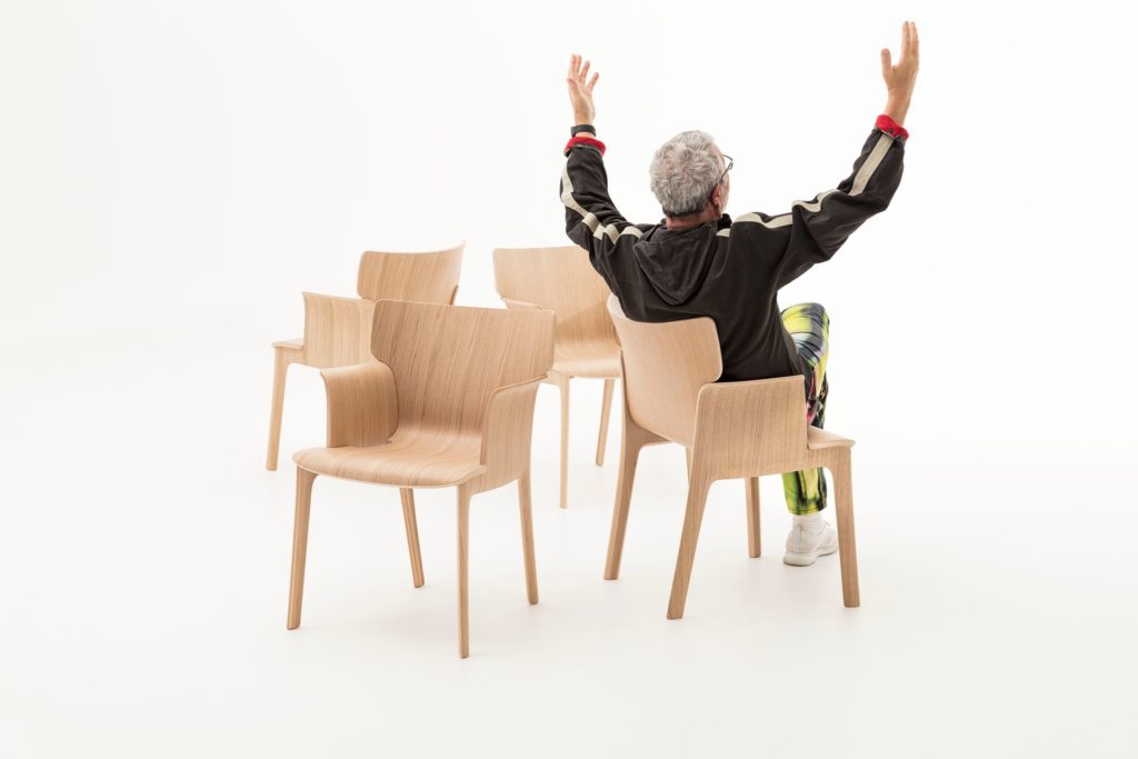 Adela Rex in oak four chairs with Starck sitting in one with arms raised view of his back 