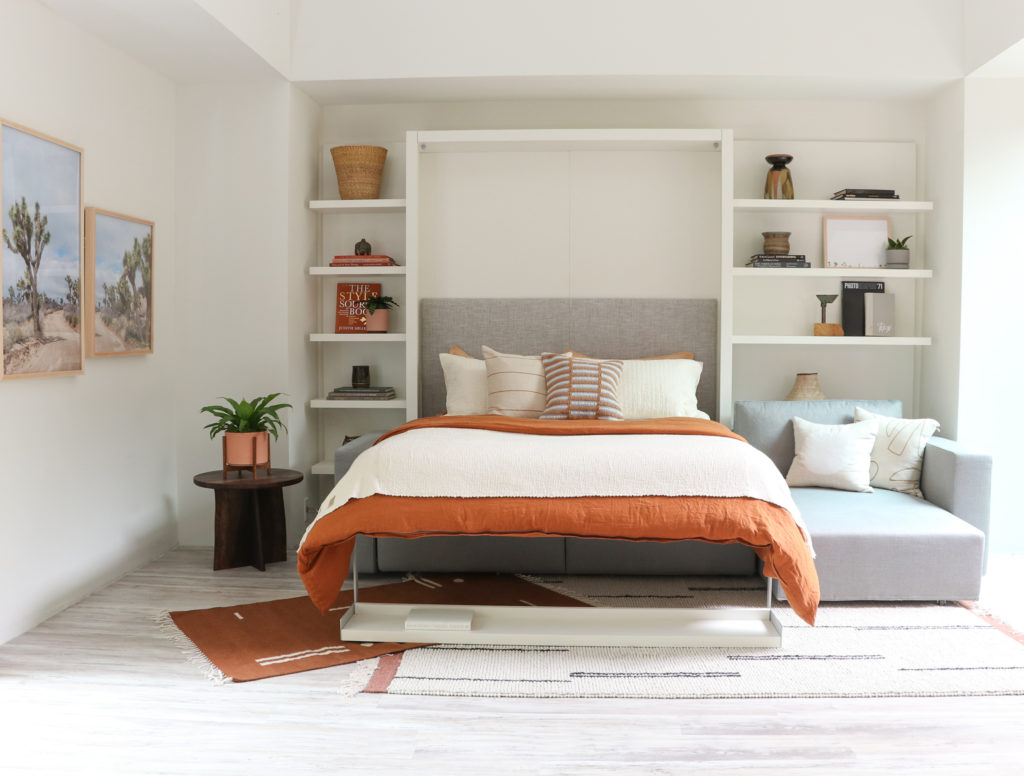 Swing as bed with orange comforter and gray upholstery on sofa with white shelves behind