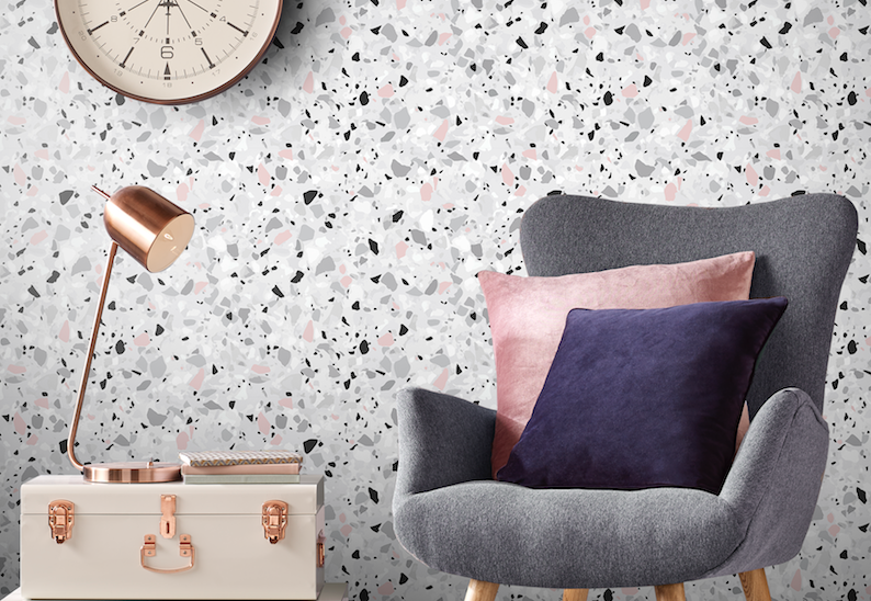 accent wall with terrazzo-inspired wallpaper with pink accents with gray upholstered chair, lamp, and antique luggage