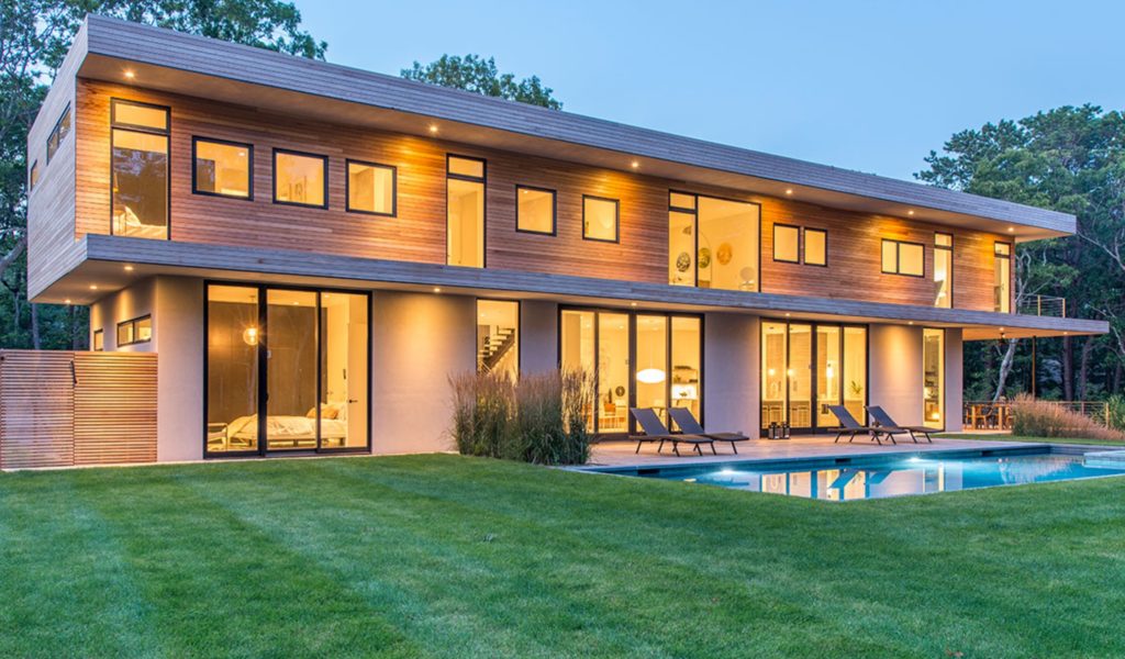 Modern Net Zero Hamptons' home with horizontal wood siding, many windows, and glass sliding doors, exterior view with outdoor pool in foreground