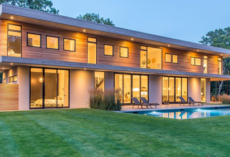Modern Net Zero Hamptons' home with horizontal wood siding and many windows and glass sliding doors, exterior view with outdoor pool in foreground