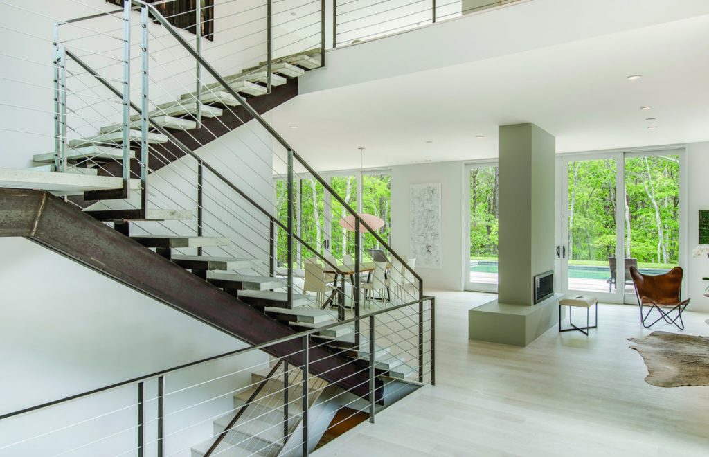 Modern Net Zero Hamptons' home interior image with open staircase and fireplace pillar with view of outdoors and swimming pool. 