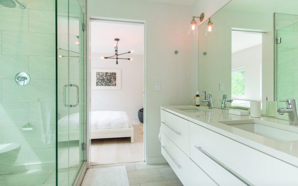 Modern Net Zero Hamptons' home interior image of bathroom with solid surface white vanity, glass shower, and partial view of bedroom beyond