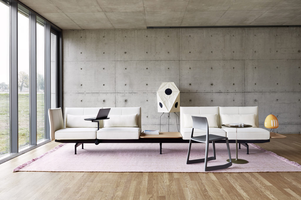 Barber & Osgerby Soft Work work system white sofa with attached rotating tables with laptops in room with concrete walls, wood floor, and large pink rug