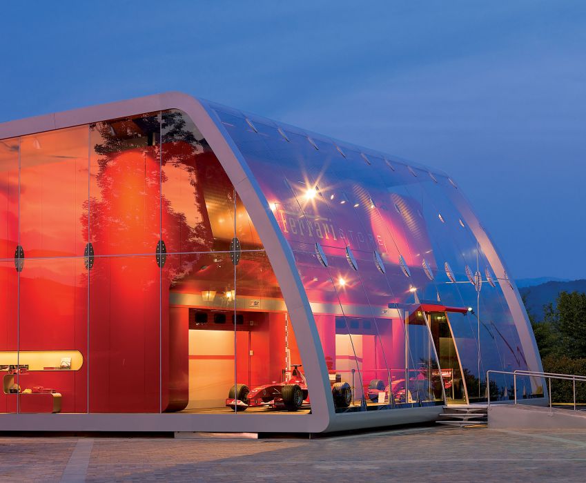 Exterior image of Ferrari Factory Store large glass walled building with red interior and Ferrari racing cars inside 