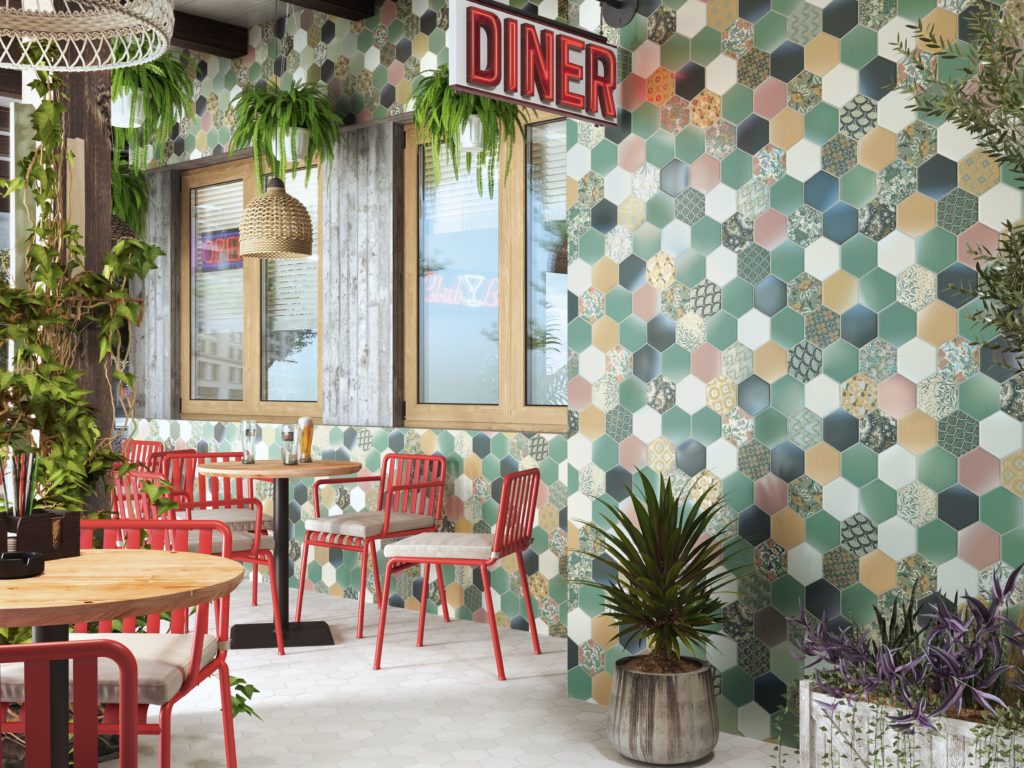 Tile of Spain 2020 designs Return to Color vibrant hexagonal tile in Mediterranean colors on outdoor wall of diner patio with wood tables and red chairs