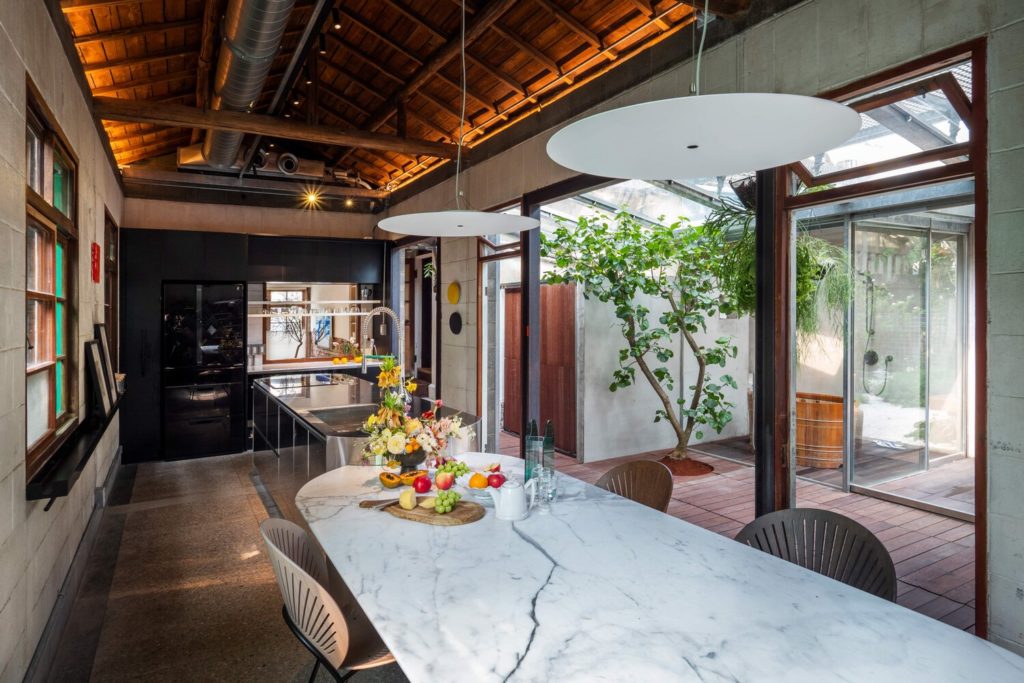 Living Lab kitchen with long marble table, stainless steel cookstove, circular overhead lights, and exposed ceiling with visible ducting and adjacent greenhouse