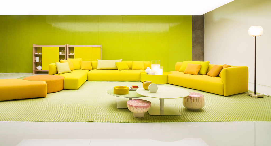 Paola Lenti Walt Seating several sofas in yellow with peach colored poof and additional seating with two separate tables in front of sofa and yellow storage unit behind