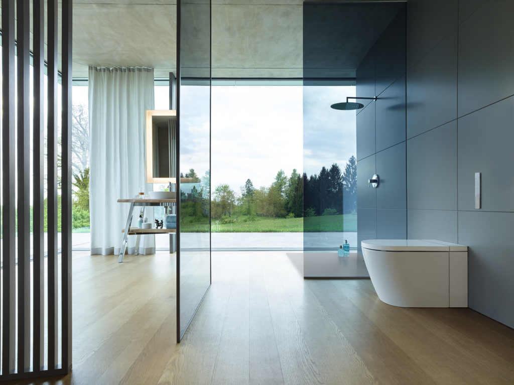 Duravit SensoWash i side view of toilet in open bathroom with glass-walled shower and exterior green hill and trees visible out window