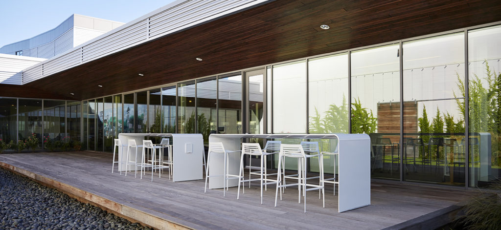 Landscape Forms' Go Outdoor Table two standing height tables in white on outdoor patio of large two-story building with large windows