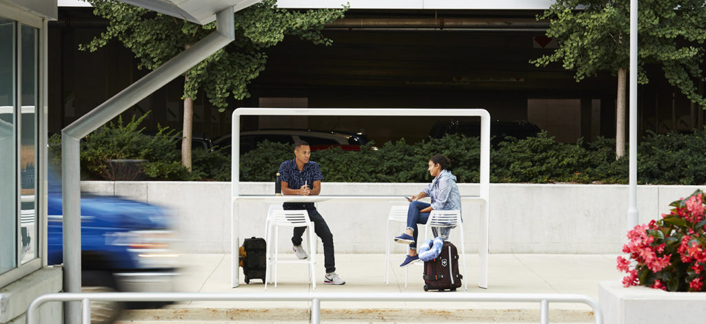Landscape Forms' Go Outdoor Table view from front standing height in white with canopy and two people talking with trees in background