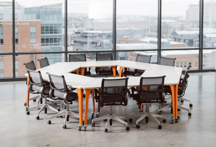 Werk Trap Table several tables with orange legs set together to form hexagonal shape surrounded by black task chairs in open room with city view