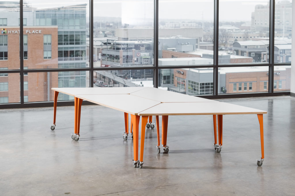 Werk Trap Table four tables with orange legs set together to form rectangular shape in open room with city view 