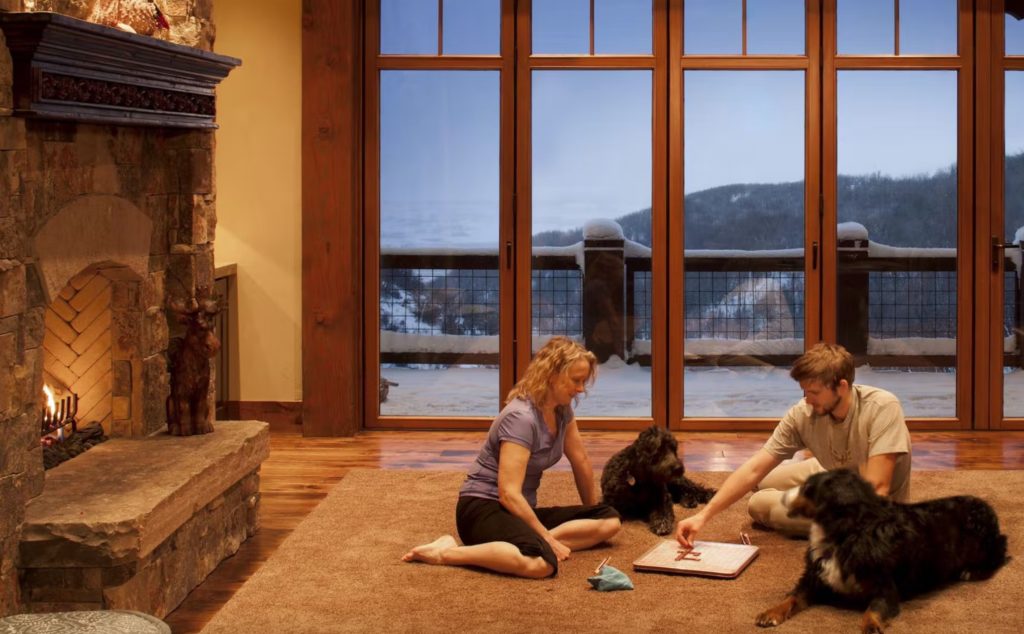 couple with dogs playing scrabble in living room of mountain house with mountains visible past glass doors 