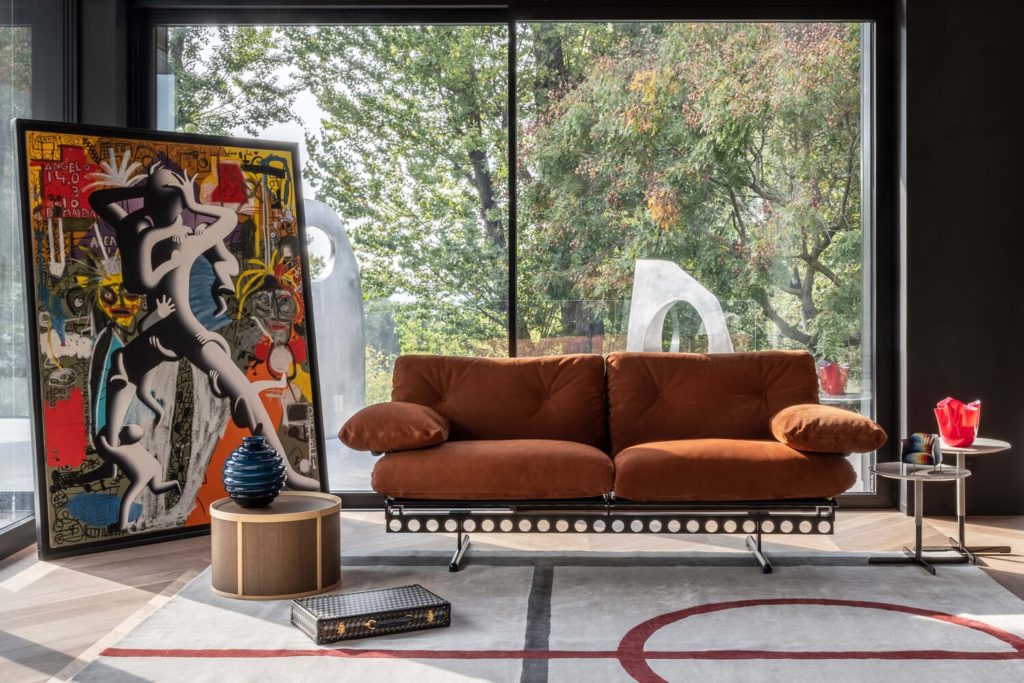 Sofa in room next to wild painting with many colors and different humanoid figures in front of large window 