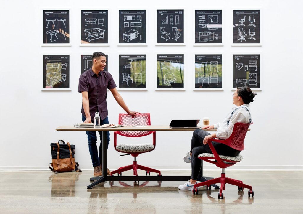 Two people in work setting with table and two red task chairs